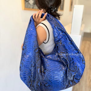 May include: A hobo-style shoulder bag featuring a vibrant blue and black snakeskin pattern with a textured, scaled surface. The bag has a slouchy, unstructured silhouette and includes the text Danastribagya printed in white on the front.