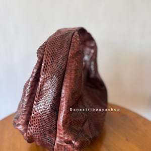 Peut inclure: Un sac &agrave; main marron rouge&acirc;tre avec un motif peau de serpent. Le sac est fait d'un mat&eacute;riau textur&eacute; et est pos&eacute; sur une surface en bois. Le texte "Danastribagyashop" est visible sur le sac.
