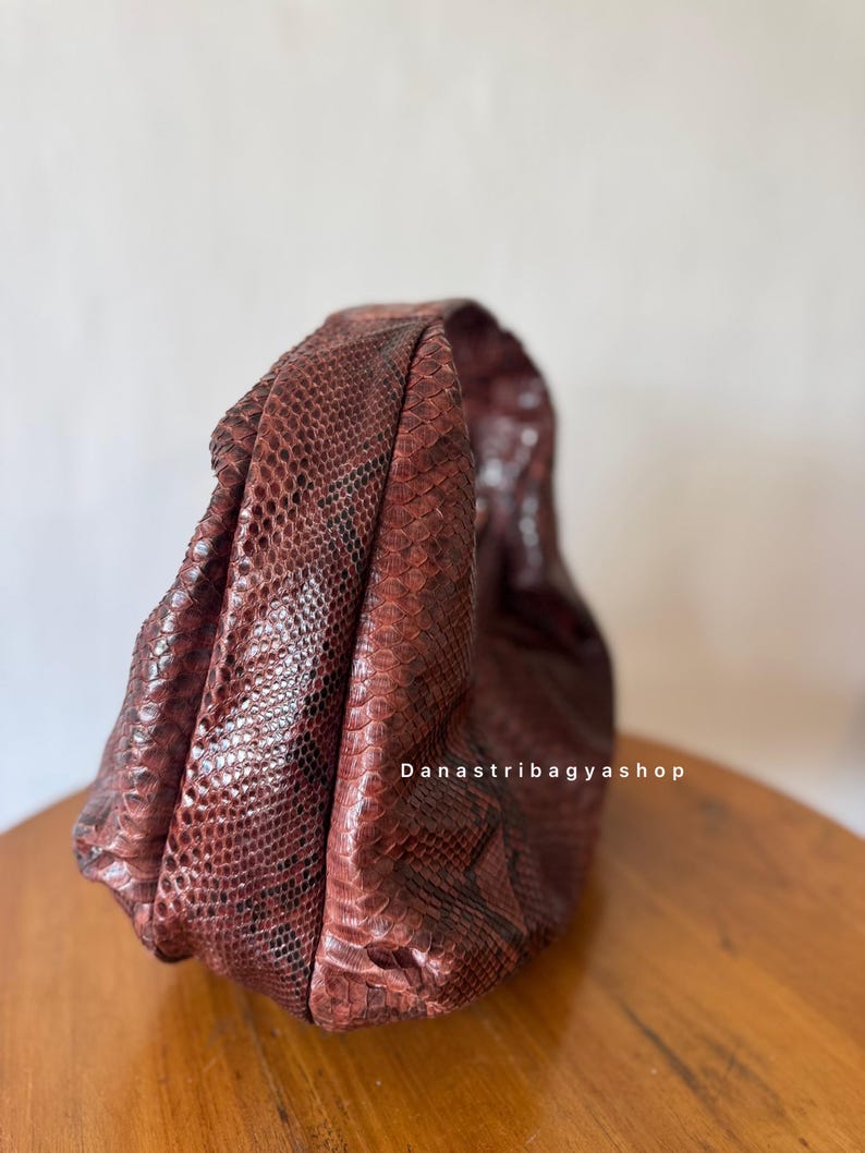 Peut inclure: Un sac &agrave; main marron rouge&acirc;tre avec un motif peau de serpent. Le sac est fait d'un mat&eacute;riau textur&eacute; et est pos&eacute; sur une surface en bois. Le texte "Danastribagyashop" est visible sur le sac.