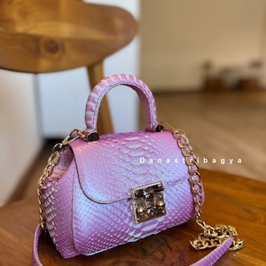 May include: A small, iridescent pink handbag with a textured snakeskin pattern. It features a top handle, a gold chain strap, and a gold clasp closure. The bag is sitting on a wooden surface.