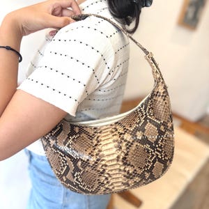May include: A crescent-shaped handbag with a snakeskin pattern in shades of brown, tan, and black. The bag features a zipper closure and a matching strap. The bag is being held over the shoulder.