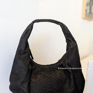 Könnte beinhalten: Eine schwarze, strukturierte Handtasche mit geschwungener Form und einem einzelnen Henkel. Die Tasche scheint aus einem Material mit schuppenartigem Muster zu bestehen, möglicherweise Leder oder einem ähnlichen Material. Die Tasche hat einen Reißverschluss.