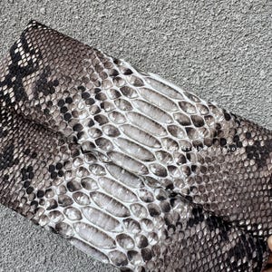 K&ouml;nnte beinhalten: Gefaltete Clutch mit Schlangenmuster in Grau-, Braun- und Wei&szlig;t&ouml;nen. Die Schuppen sind detailliert und strukturiert, wodurch ein realistisches Aussehen entsteht. Die Tasche ist rechteckig und scheint aus Leder zu sein.