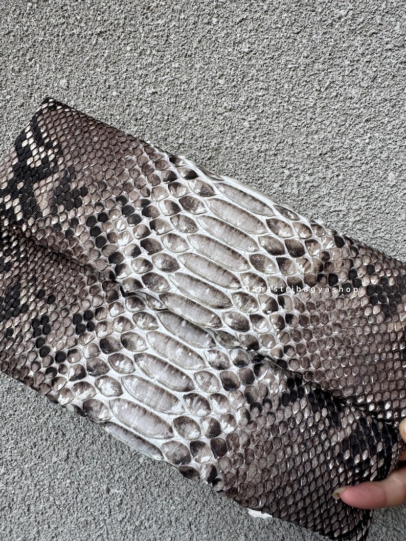K&ouml;nnte beinhalten: Gefaltete Clutch mit Schlangenmuster in Grau-, Braun- und Wei&szlig;t&ouml;nen. Die Schuppen sind detailliert und strukturiert, wodurch ein realistisches Aussehen entsteht. Die Tasche ist rechteckig und scheint aus Leder zu sein.