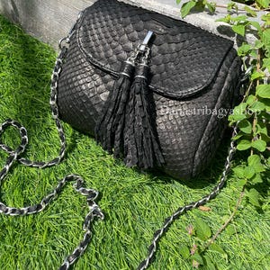 Op de afbeelding: Zwarte crossbodytas met een textuur met schubbenpatroon. De tas heeft een klepsluiting, een zilveren kettingriem en twee zwarte kwastjes. De tas ligt op groen gras.