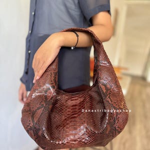 May include: A brown leather handbag with a snakeskin pattern. The bag has a curved shape and a single handle. The text "Danastribagyshop" is visible on the bag.