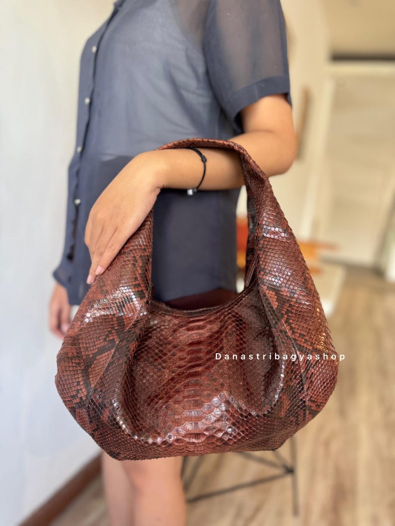 Peut inclure: Un sac &agrave; main en cuir marron avec un motif peau de serpent. Le sac a une forme incurv&eacute;e et une seule poign&eacute;e. Le texte "Danastribagyshop" est visible sur le sac.