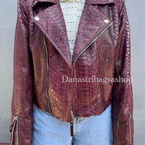 Pode incluir: Uma jaqueta de couro bordô com padrão de pele de cobra. A jaqueta tem fecho de zíper e gola. A jaqueta é curta e tem mangas compridas.