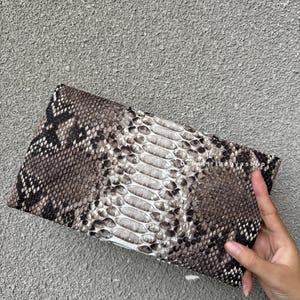 K&ouml;nnte beinhalten: Eine rechteckige Clutch mit Schlangenmuster in Grau-, Braun- und Wei&szlig;t&ouml;nen. Die Clutch hat ein strukturiertes Aussehen mit einem zentralen Feld aus helleren Schuppen. Der Shop-Name "DabaTribagyashop" ist oben rechts sichtbar.
