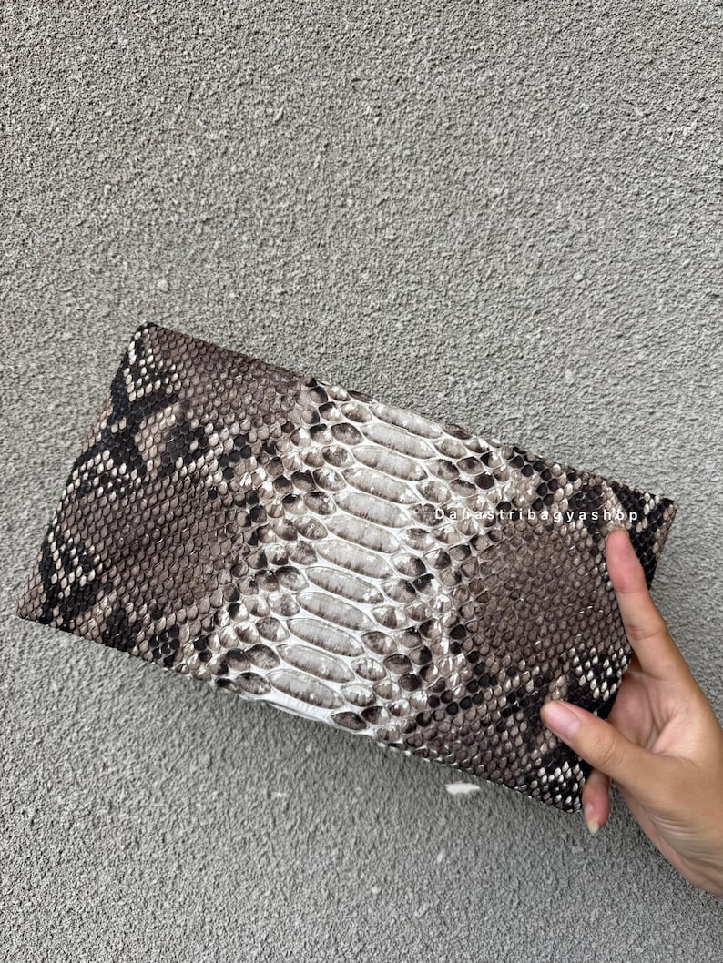 K&ouml;nnte beinhalten: Eine rechteckige Clutch mit Schlangenmuster in Grau-, Braun- und Wei&szlig;t&ouml;nen. Die Clutch hat ein strukturiertes Aussehen mit einem zentralen Feld aus helleren Schuppen. Der Shop-Name "DabaTribagyashop" ist oben rechts sichtbar.