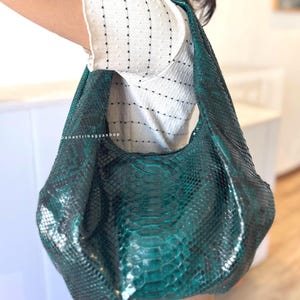 May include: A teal green leather shoulder bag with a snakeskin pattern. The bag has a large, curved shape and a single shoulder strap. The text "danastribagyashop" is visible on the bag.