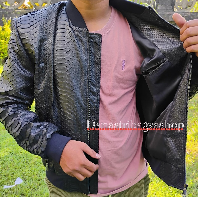 May include: A black leather bomber jacket with a snakeskin pattern. The jacket is unzipped, showing the black lining. The jacket has a zipper closure and a ribbed collar.