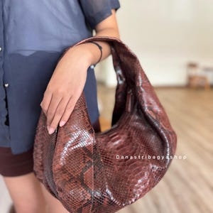 Peut inclure: Un sac &agrave; bandouli&egrave;re en cuir marron avec un motif peau de serpent. Le sac est tenu par une personne. Le sac a une forme incurv&eacute;e et une seule sangle. Le texte "Danastribagyashop" est visible sur le sac.