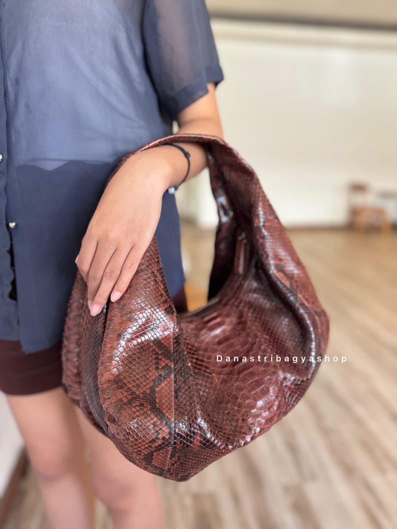 Peut inclure: Un sac &agrave; bandouli&egrave;re en cuir marron avec un motif peau de serpent. Le sac est tenu par une personne. Le sac a une forme incurv&eacute;e et une seule sangle. Le texte "Danastribagyashop" est visible sur le sac.