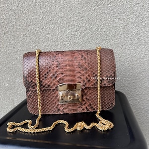 May include: A brown, rectangular handbag with a gold chain strap and a gold clasp. The bag has a snakeskin pattern and is sitting on a black surface. The bag is a small, crossbody style.