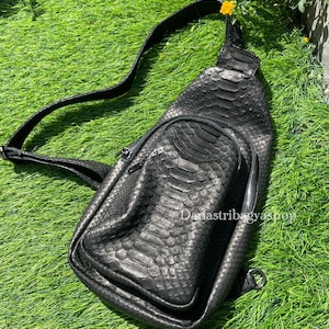 Pode incluir: Bolsa transversal preta com um padrão texturizado semelhante à pele de cobra. A bolsa possui um compartimento principal com zíper e uma alça ajustável. A bolsa é feita de um material escuro.