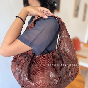 Peut inclure: Un sac &agrave; bandouli&egrave;re marron et rouge &agrave; motif peau de serpent. Le sac a une forme de croissant et un design souple et ample. Le texte "Danastribagyashop" est visible sur le sac.