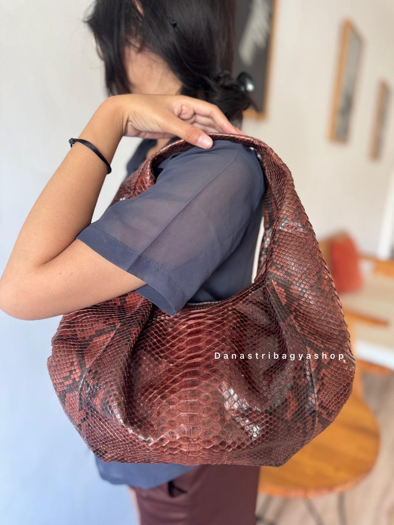 Peut inclure: Un sac &agrave; bandouli&egrave;re marron et rouge &agrave; motif peau de serpent. Le sac a une forme de croissant et un design souple et ample. Le texte "Danastribagyashop" est visible sur le sac.