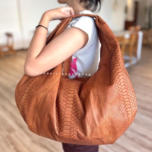 May include: A large, brown leather hobo bag with a snakeskin texture. The bag has a wide, curved strap and a slouchy design. The bag is carried over the shoulder. The bag is a warm, earthy tone.