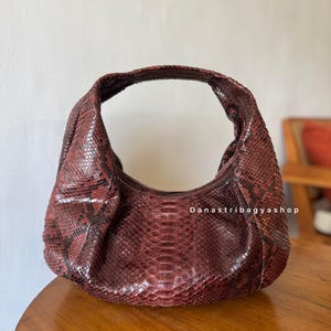 Peut inclure: Un sac &agrave; main en forme de croissant de couleur bordeaux avec un motif peau de serpent. Le sac a une seule poign&eacute;e et une forme arrondie. La texture de la peau de serpent est visible, et le sac semble &ecirc;tre en cuir. Le texte "Danastribagyashop" est visible.