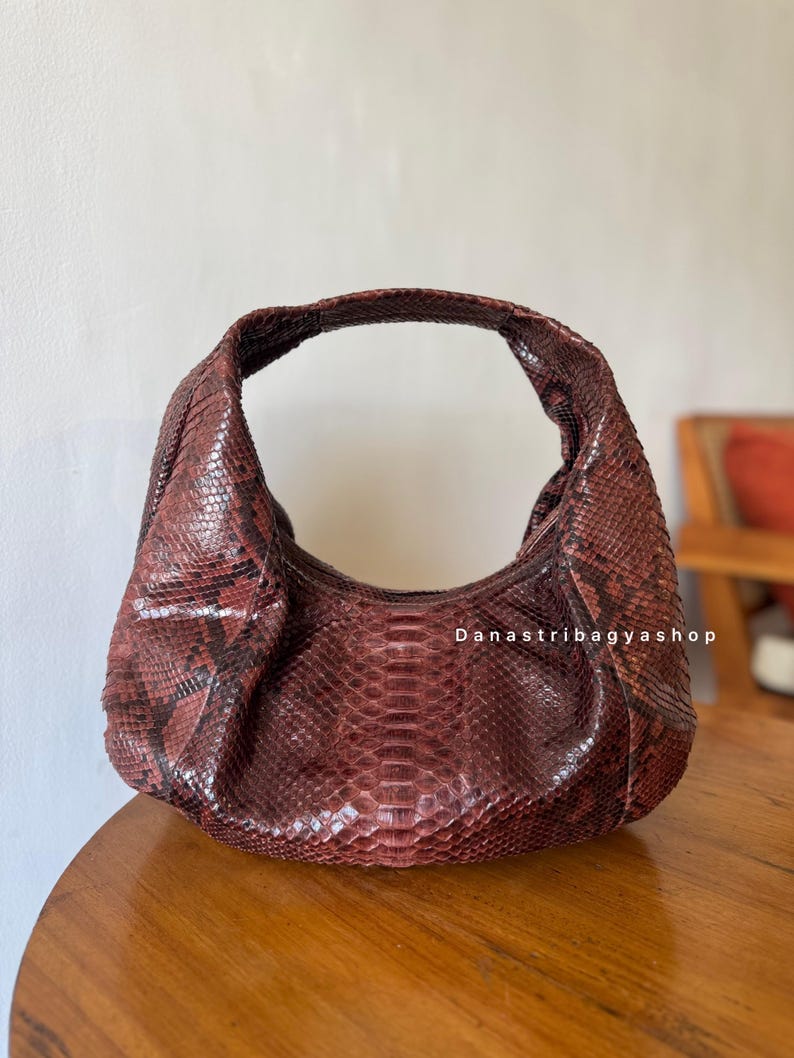 Peut inclure: Un sac &agrave; main en forme de croissant de couleur bordeaux avec un motif peau de serpent. Le sac a une seule poign&eacute;e et une forme arrondie. La texture de la peau de serpent est visible, et le sac semble &ecirc;tre en cuir. Le texte "Danastribagyashop" est visible.