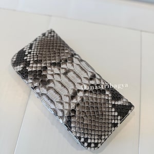 May include: A rectangular wallet featuring a textured snakeskin pattern in shades of gray, black, and white. The surface displays a realistic scale design with a gradient effect. The text Danastribagya is printed in white on the center of the wallet.