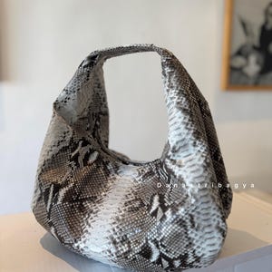 May include: A hobo-style handbag featuring a textured snakeskin pattern in shades of white, grey, and black. The bag has a single integrated handle and a curved, slouchy silhouette. The text 'Danastribagya' is printed in white on the side.