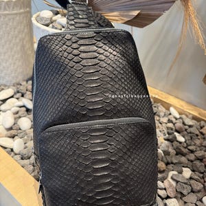 Könnte beinhalten: Schwarze Leder-Umhängetasche mit Schlangenmuster. Die Tasche hat ein Reißverschluss-Hauptfach und eine Vordertasche. Der Riemen ist verstellbar. Die Tasche liegt auf einer Oberfläche aus kleinen Steinen.