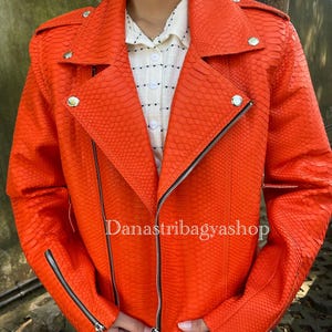 Pode incluir: Casaco de couro laranja vibrante com textura de pele de cobra, com fecho de correr assimétrico, detalhes prateados e gola de lapela. O casaco tem um corte ajustado e o texto "Danastribagyashop" é visível.