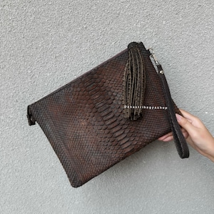 Peut inclure: Une pochette en cuir marron avec un motif peau de serpent. La pochette est dotée d'une dragonne et d'un pompon décoratif. L'image est prise sur un fond gris. Le texte "danastribagyashop" est visible sur l'image.