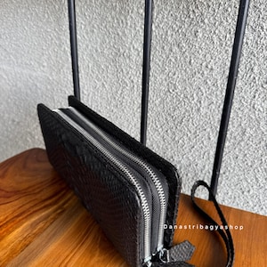 May include: Black wallet with a textured pattern and two silver zips. The wallet has a wrist strap and is displayed on a wooden surface. The wallet is a long, rectangular shape.