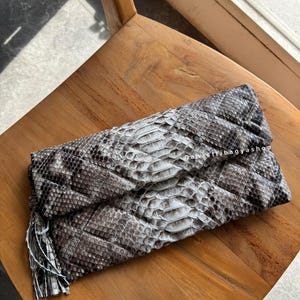 Puede incluir: Bolso de mano de piel de serpiente en tonos grises y blancos, con detalle de borla. El bolso está doblado y descansa sobre una silla de madera. El texto "Danastribagyshop" es visible.