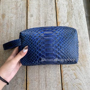 Pode incluir: Uma clutch retangular azul com um padrão de pele de cobra texturizado. A clutch tem uma pequena alça lateral. O texto "Danastribagyashop" está impresso na frente.