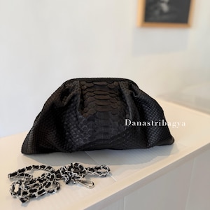 May include: A black textured snakeskin-patterned clutch bag with a gathered frame closure. The bag features a detachable silver-tone chain strap with black woven detailing. The text Danastribagya is printed in white on the right side of the bag.