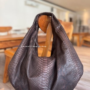 Puede incluir: Un bolso de hombro de cuero marrón con un estampado de piel de serpiente. El bolso está colgado de una silla de madera. El texto "Danastribagyashop" es visible en el bolso.
