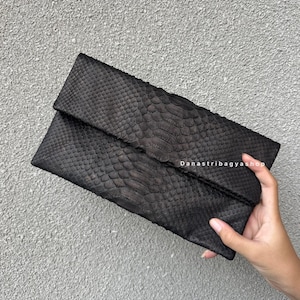 May include: A black clutch with a snakeskin pattern. The rectangular bag has a fold-over flap and is held by a hand. The text "Danastribagyashop" is visible on the bag.