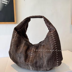 Op de afbeelding: Een bruine handtas met slangenleertextuur en een gebogen, halvemaanvorm. De tas heeft een enkele handgreep en een ruim interieur. De tekst "Danastribagyshop" is zichtbaar op de zijkant van de tas. De tas staat op een wit oppervlak.