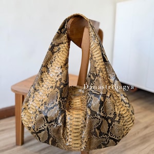 May include: A hobo-style handbag featuring a textured snakeskin pattern in shades of tan, brown, and black. The bag has a single integrated shoulder strap and a slouchy, rounded silhouette. The text Danastribagya is printed on the front surface.