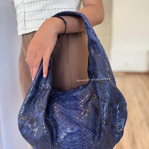 May include: A blue leather handbag with a snakeskin pattern. The bag has a rounded shape and a single shoulder strap. The bag is held by a person. The text "Danastribagyashop" is visible on the bag.