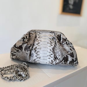 May include: A clutch bag featuring a gray, white, and black snakeskin pattern with a gathered, pouch-like silhouette. The bag includes a detachable silver-toned metal chain strap and displays the text Danstibagya on the side.