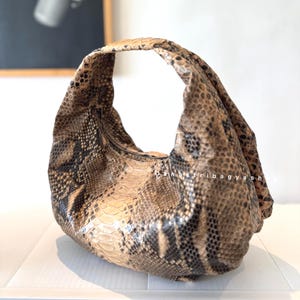 May include: A hobo-style handbag featuring a textured snakeskin pattern in shades of brown, tan, and black. The bag has a single integrated handle and a slouchy, rounded silhouette. The text "danastribagyshop" is printed in white on the side.