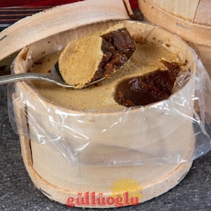 May include: A wooden container filled with a light brown, creamy dessert, with a darker brown, textured filling. A spoon holds a portion of the dessert, revealing its layered texture. The container is lined with clear plastic.