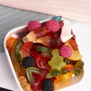 May include: A white square bowl filled with a colorful assortment of gummy candies. The candies include shapes like bottles, berries, and trees in shades of red, yellow, green, and orange. Some candies are topped with small, pink, round candies.