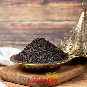 May include: A bronze bowl filled with dark, granular spice, possibly sumac, is on a wooden board. An ornate, decorative lid is nearby. The image has a warm tone, suggesting a culinary theme. The logo "güllüoğlu" is visible.