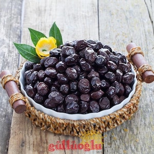 Può includere: Una ciotola bianca piena di olive scure e lucide, presentata su un cestino intrecciato. La ciotola è adornata da un fiore giallo e foglie verdi. I manici in legno del cestino sono visibili e lo sfondo è una superficie di legno.
