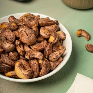 Puede incluir: Un cuenco blanco lleno de anacardos tostados. Los anacardos son una mezcla de colores marrón y beige, con un aspecto ligeramente texturizado. Algunos anacardos están esparcidos fuera del cuenco sobre una superficie verde. La imagen está bien iluminada.