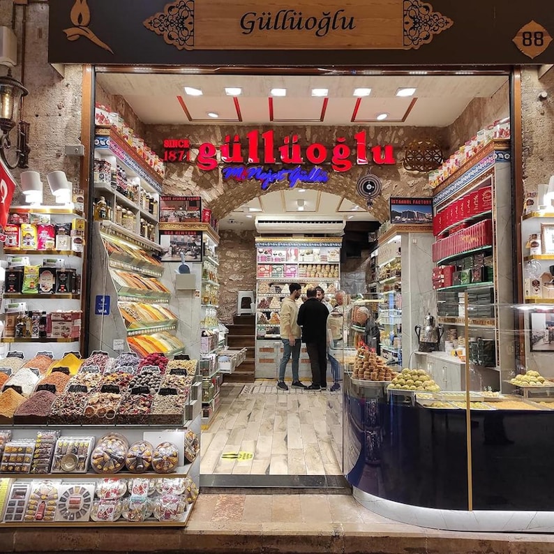 May include: A shop front displaying a range of sweets and spices. The sign above reads "G&uuml;ll&uuml;oğlu" in red neon. Shelves are filled with packaged goods, and customers are seen inside the store, browsing the selection.