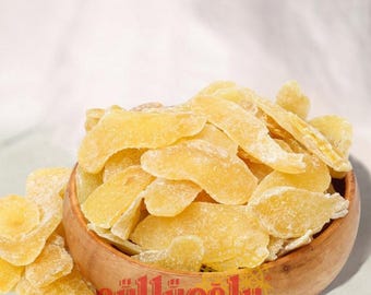 May include: A wooden bowl filled with candied ginger slices. The ginger is a light yellow color and has a slightly crystallized surface. The bowl is sitting on a white surface. The text "güllüoğlu 1871" is visible on the bowl.
