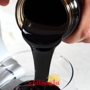 May include: A dark, viscous liquid being poured from a glass jar into a clear glass. The liquid is a deep, rich black colour, and the jar has a gold-coloured rim. The bottom of the glass has a logo with the text "gall&uuml;oğlu".