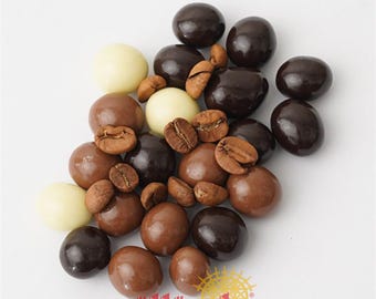 May include: An assortment of chocolate-covered candies in various shades of brown and white, along with whole coffee beans. The candies are arranged on a white surface, with the brand name "güllüoğlu" and the year "1871" visible in the lower right corner.